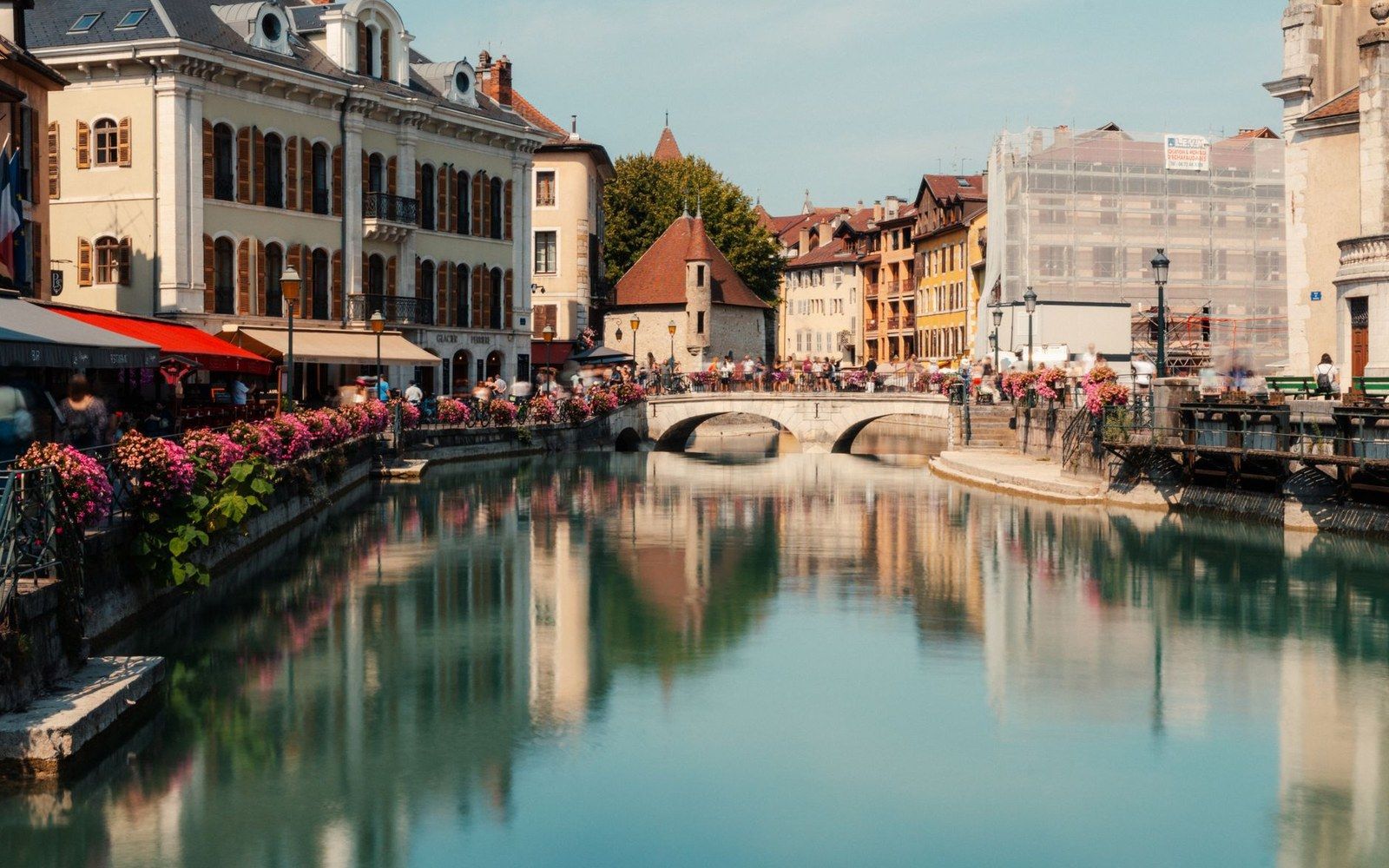 Stone bridge crosses a calm canal lined with flower-draped buildings and outdoor cafes